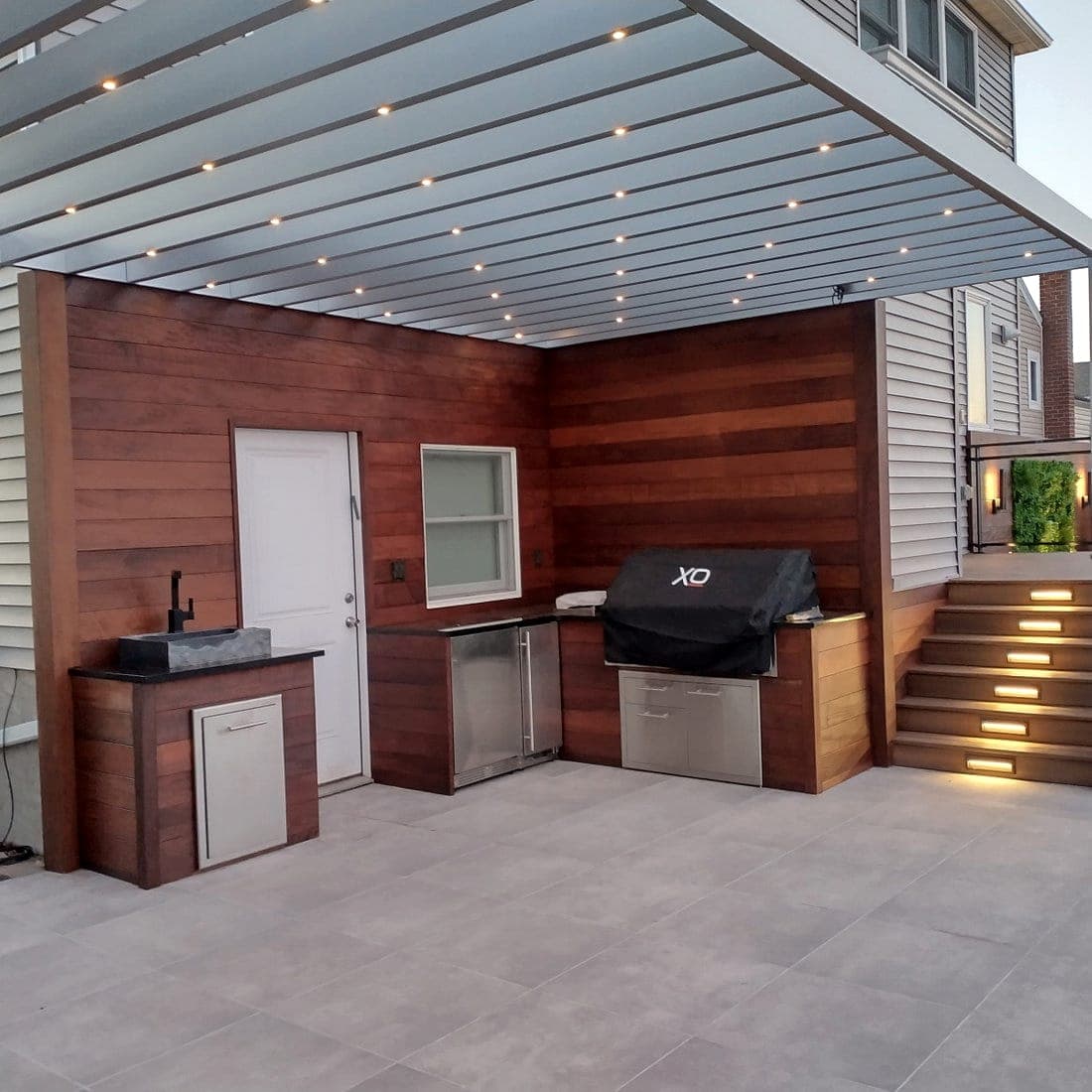 Outdoor kitchen and bar under a louvered pergola, lit at dusk with downlights